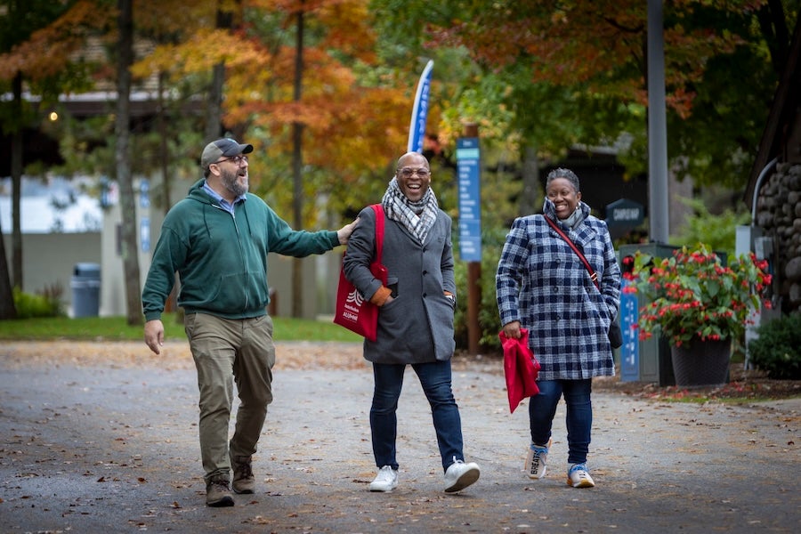 Three Interlochen alumni walking on along a tree-lined path with colorful autumn foliage.