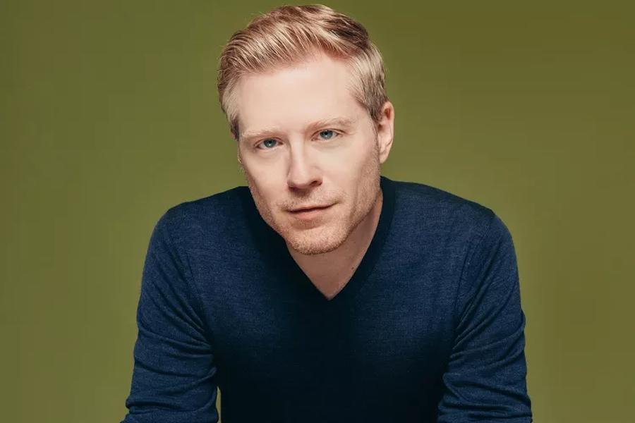 Anthony Rapp is phtoographed wearing a darl blue swather in front of a gren background. 