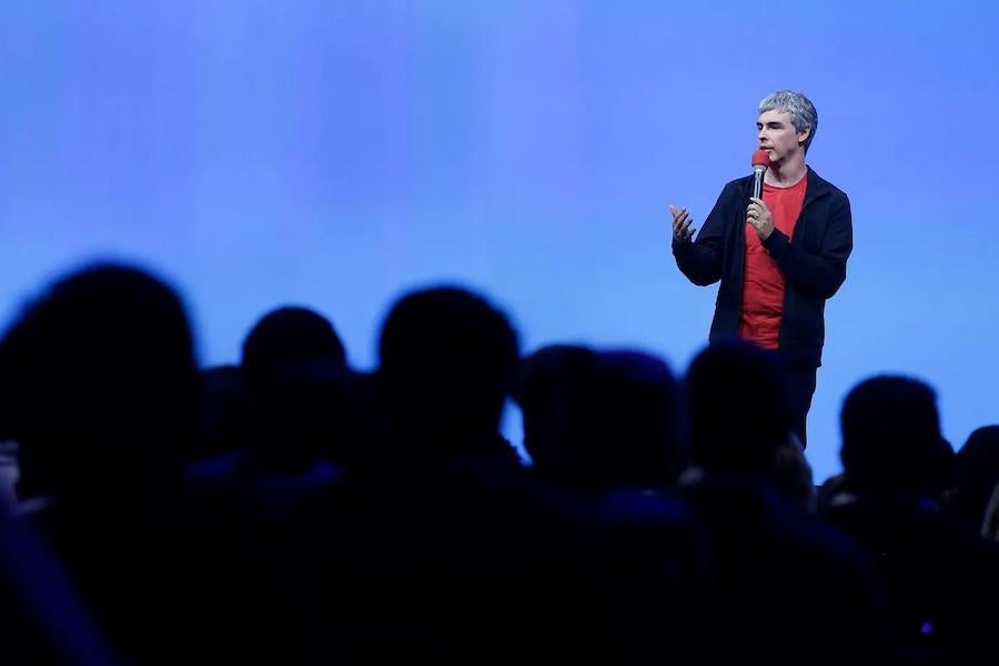 Larry Page giving a speech on a stage with a microphone in hand, gesturing to a silhouetted audience in the foreground with a blue background. 