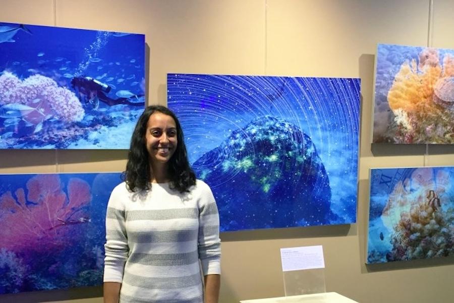  A woman in a striped sweater smiling at the camera in front of a wall displaying colorful underwater photographs of coral.