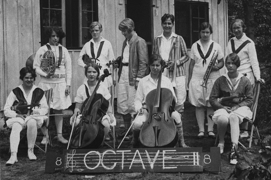 A group of young women wearing white uniforms and holding musical instruments gather in front of a wooden cabin for a group photo. 