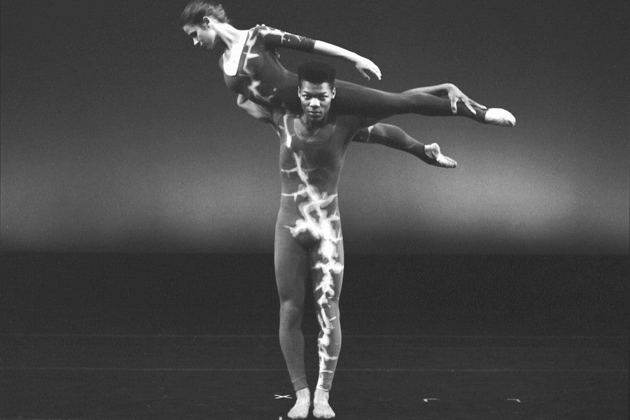 Two ballet dancers on stage, with the female dancer balanced horizontally on the male dancer's shoulder. Both wear patterned costumes.