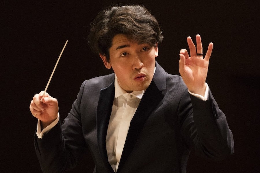 Person in a dark suit conducting with a baton, expressive gesture with the right hand, against a dark background.