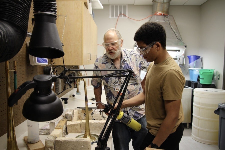 A student welds a trumpet while Ken Larson watches.