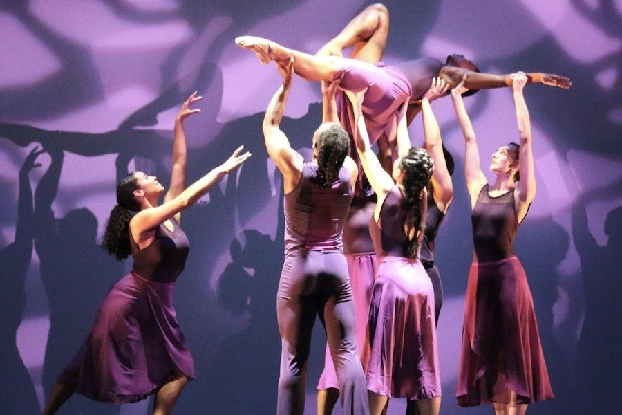 A group of dancers dressed in purple lift a dancer in the air.