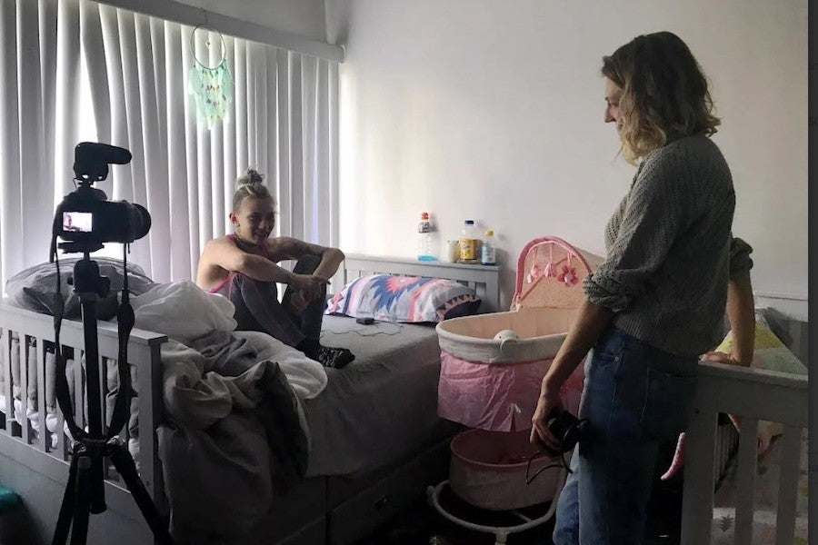 Claire Collins, standing, converses with Mckenzie Trahan in a bedroom with a camera and baby bassinet visible.