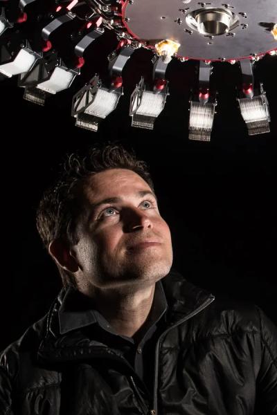 Aaron Parness, wearing a black jacket, looks upward at a circular array of robotic arms in  a dark room. 
