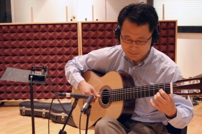 A man wearing headphones and glasses plays a guitar in front of a microphone.