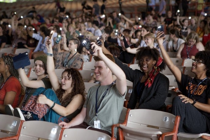 A group of students sitting in an auditorium smile and raise their phones with flashlights turned on.