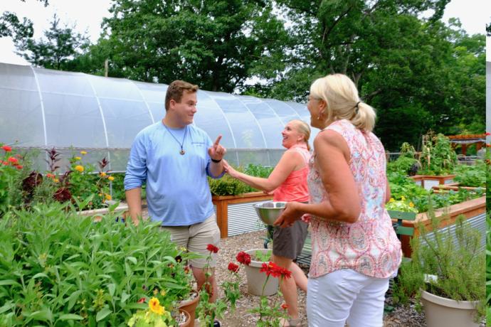 An instructor works with two adult students in a garden with a greenhouse in the background.