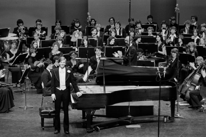 A pianist standing beside a grand piano in front of an orchestra in a black-and-white photo.
