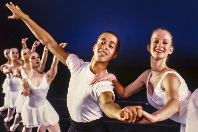 Male and female dancers smiling and holding hands in a staged performance with other dancers in the background.