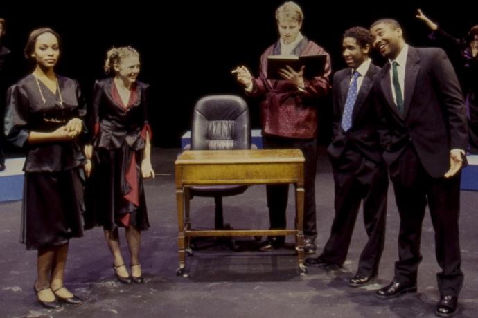 A theater performance with two women and three men on stage, standing around a wooden desk. A tall man in a lounge jacket jacket is reading a book books, while the two men on the right smile and address the two women on the left. 