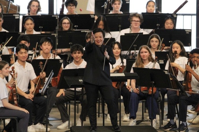 A smiling conductor, surrounded by young musicians, gestures towards the viewer.