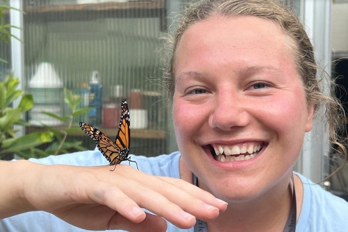 A smiling woman poses with a monarch butterfly on her hand.