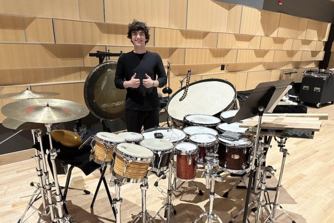 A dark-haired male student gives a thumbs up signal behind a large drum set.