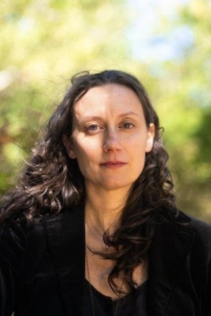 A woman with dark curly hair wearing a black shirt gazes at the camera.