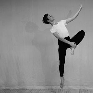  A dancer in black pants and a white shirt leaps with one leg extended downward and  the opposite foot touched to the inner thigh, extending one arm elegantly upward, captured in black and white.