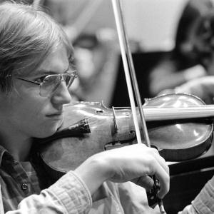 Young musician in glasses playing a violin focusedly, in a black and white photo, with another person faintly visible in the background.