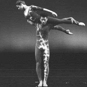 Two ballet dancers on stage, with the female dancer balanced horizontally on the male dancer's shoulder. Both wear patterned costumes.