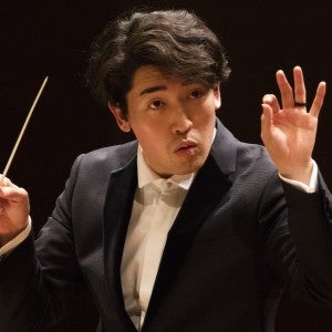 Person in a dark suit conducting with a baton, expressive gesture with the right hand, against a dark background.