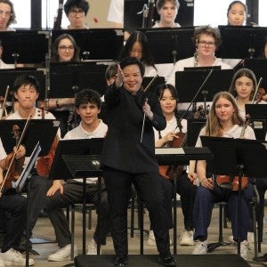 A smiling conductor, surrounded by young musicians, gestures towards the viewer.