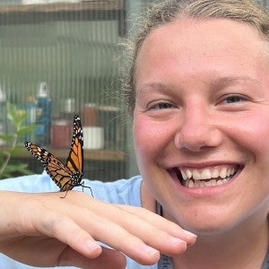 A smiling woman poses with a monarch butterfly on her hand.