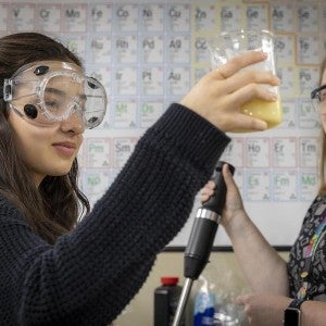 A student wearing safety goggles holds up a beaker filled with yellow liquid while another student watches.