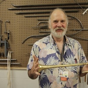 Ken Larson stands with a trumpet bell in his hands in front of a wall covered with metal tools.