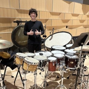 A dark-haired male student gives a thumbs up signal behind a large drum set.