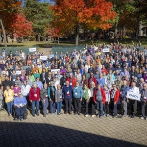 A large group of excited people gather in an outdoor auditorium, holding class year signs.