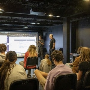 Two instructors gesture towards a screen in front of a group of seated students.