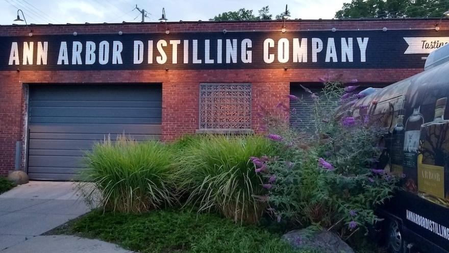 Brick building labeled "ANN ARBOR DISTILLING COMPANY" with garage doors, ornamental grasses, and a trailer with an advertisement.