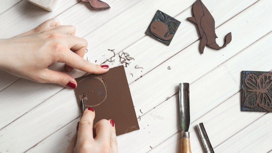 Hands carving a design into a linoleum block with tools on a wooden table.